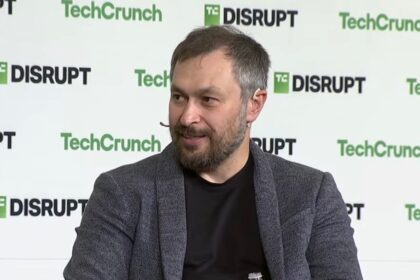 A bearded man grins against a TechCrunch-branded backdrop